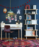 Oeuf NYC Vertical Mini Library met kinderboeken en decoratie naast een bureau tegen een donkerblauwe muur