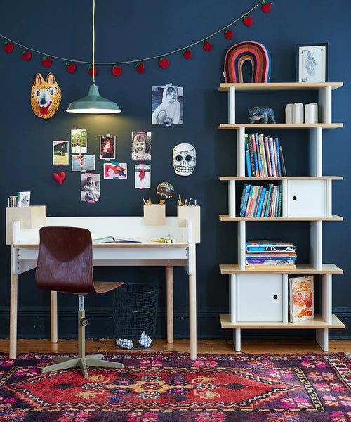 Oeuf NYC Vertical Mini Library met kinderboeken en decoratie naast een bureau tegen een donkerblauwe muur