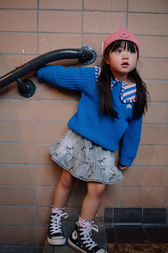 Meisje draagt de koraalroze Club Chaos beanie van Sproet & Sprout gecombineerd met een blauwe trui uit dezelfde collectie.